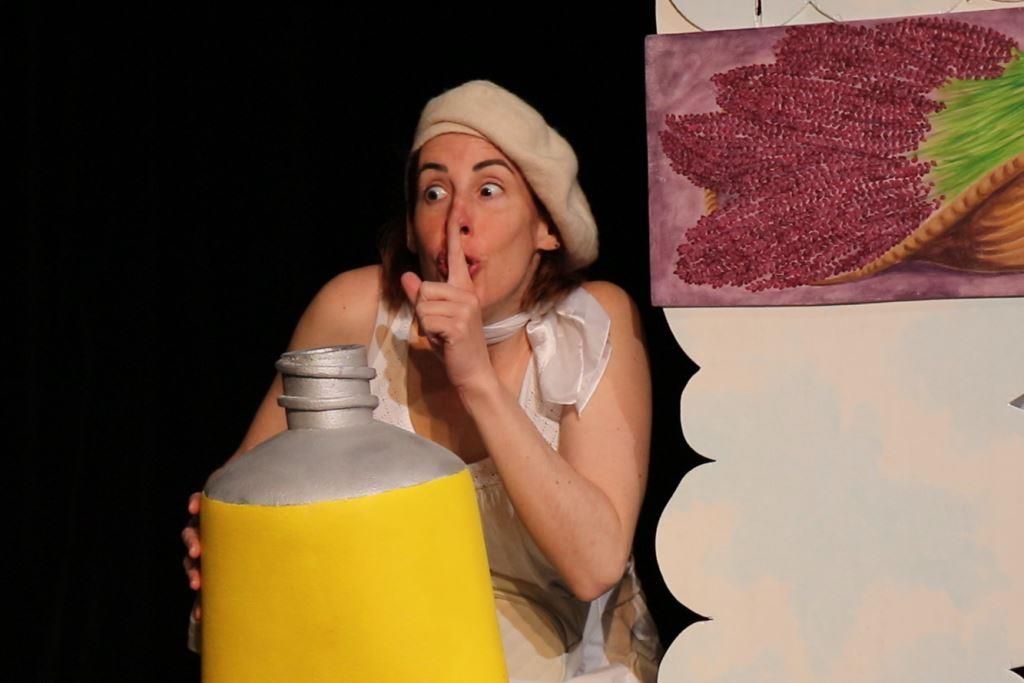 La comédienne au béret blanc, un doigt sur la bouche, tient un tube de peinture jaune géant devant un tableau de fleurs roses.