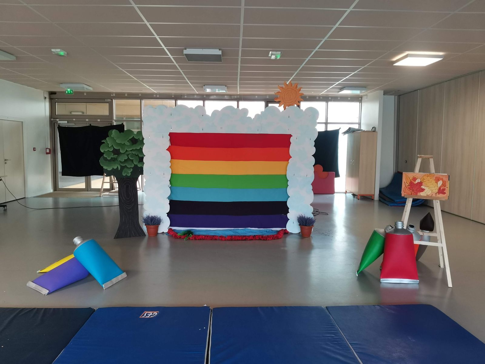 Le décor monté dans une salle ordinaire : un arc-en-ciel de tissu encadré de nuages découpés, un arbre en carton, un chevalet et des tubes de peinture géants posés au sol.