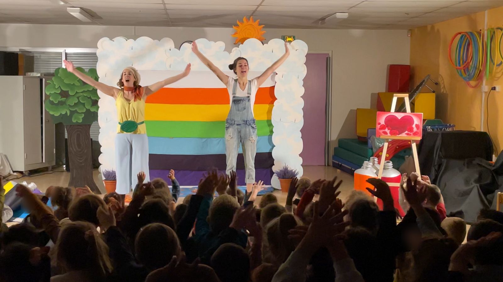Une classe assise au sol dans la salle de motricité d’une école, mains levées vers les deux comédiennes qui chantent bras ouverts devant l’arc-en-ciel de tissu.